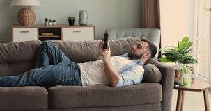 Relaxed millennial guy lie on back on soft comfortable couch at living room spend lazy weekend watching video on mobile phone screen. Calm young man rest use cellphone to check email read news online
