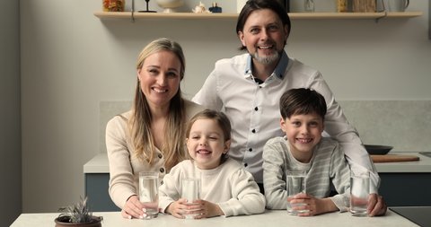 Happy couple and little children holds glasses with quality pure still water pose in kitchen look at camera. Healthy life habit, home water purification system, health-care, natural hydration concept
