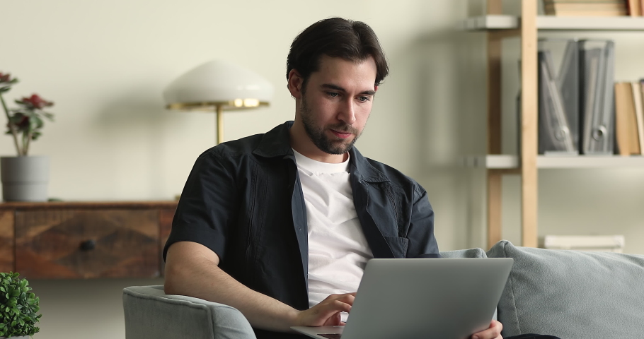 Young handsome man sit on sofa working or shopping use laptop, search information on internet, websurfing relax at home, do telecommute from homeoffice. Modern wireless tech usage, e business concept Royalty-Free Stock Footage #1085054404