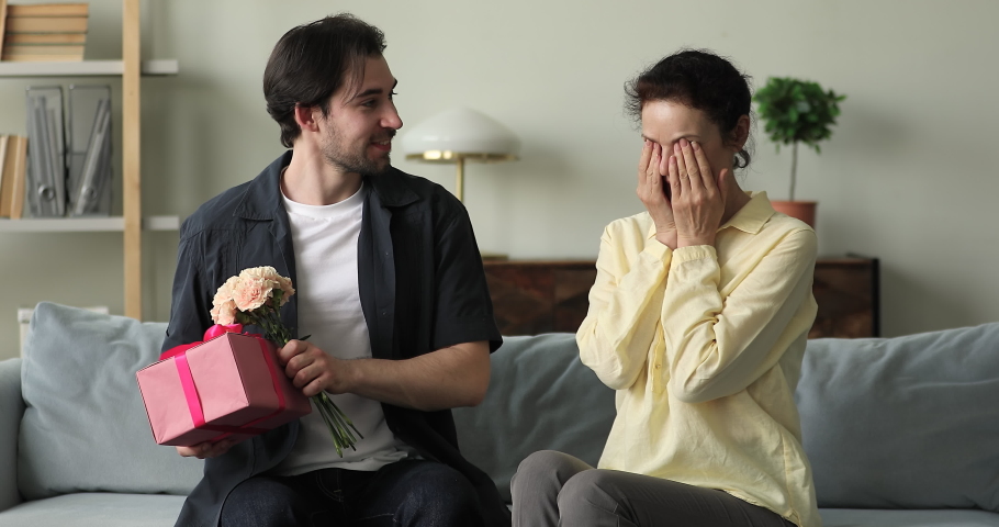 Older woman sit on sofa, cover eyes with palms wait surprise prepared from grown up son, get flowers, gift box feels happy hugs him. Mother Day, life event celebration, birthday congratulation concept Royalty-Free Stock Footage #1085054422