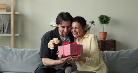 Older woman sit on sofa, cover eyes with palms wait surprise prepared from grown up son, get flowers, gift box feels happy hugs him. Mother Day, life event celebration, birthday congratulation concept