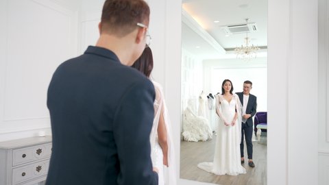 4K Asian LGBTQ guy bridal shop owner helping female bride customer choosing and trying on wedding gown at wedding studio. Small business entrepreneur wedding planner and marriage ceremony concept