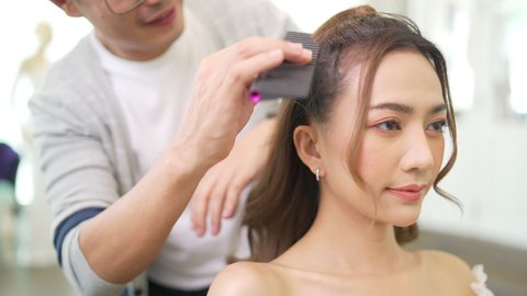 4K Asian LGBTQ guy hairdresser brushing hair to young beautiful woman bride in wedding dress at wedding studio. Small business entrepreneur bridal shop owner and  beauty salon hair stylist concept.