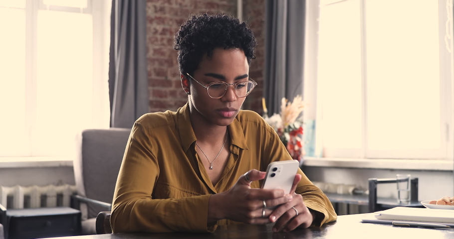 Young African woman sit at table at home office holds cell phone smile text message chat with friend, search information on internet, make order, enjoy easy e-commerce services. Wireless tech concept Royalty-Free Stock Footage #1085056226