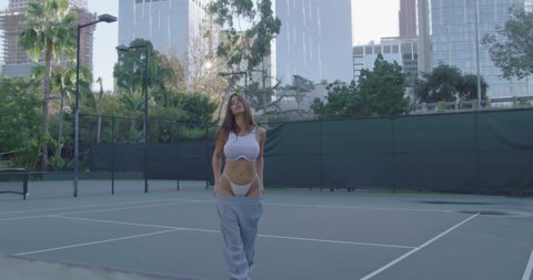 A beautiful sexy girl goes and shows her ass in a white bikini on a tennis court on red camera, slow motion at sunset