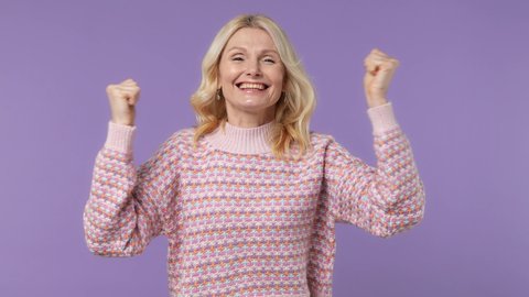 Excited jubilant happy elderly blonde woman lady 40s years old wears warm shirt doing winner gesture celebrate clenching fists say yes isolated on plain pastel light purple background studio portrait