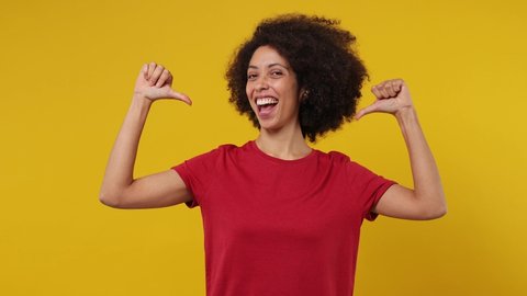 Confident blithesome charming young black woman 20s years old wears red t-shirt pointing fingers on herself blinking showing thumb up like gesture isolated on plain yellow background studio portrait