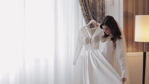 Bride in lingerie dancing with her wedding dress. White boudoir dress. Morning preparations indoors. Luxury bride with hairstyle and makeup in a hotel or apartment. Woman in white night gown and veil