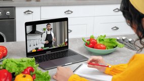 Man food blogger in computer screen tells teaches housewife online video call webcam laptop, slices cucumber. Woman in kitchen study remote culinary lesson listen chef teacher, writes in notebook