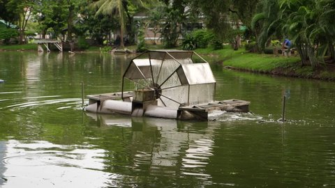 Floating aerator machine work on green water of lake in Lumphini park, slow motion shot. Water turbine rotate and pour down many trickles of water. Lush tropical vegetation on background
