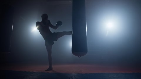 Kickboxing. Silhouette of young man training with punching bag in boxing club. Sportsman boxing in smoky studio. Male strikes with his feet. Darkness background. Sport concept. 4K, UHD