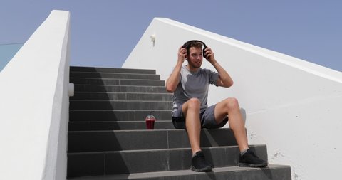 Man listening to music, audiobook or podcast mobile phone app with headphones sitting on stairs. Healthy city lifestyle person using smartphone on running break with morning red beet juice smoothie
