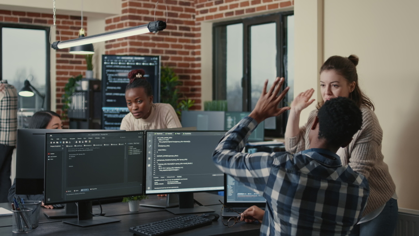 Programmer analyzing compiling code on multiple screens takes off glasses and doing high five hand gesture with colleague developer. Software developers celebrating successful coding. | Shutterstock HD Video #1085102060