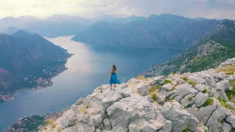 A young girl in a blue dress stands on the top of a mountain overlooking the Bay of Kotor in Montenegro. Aerial drone view 4K. Lifestyle tourism and beauty concept