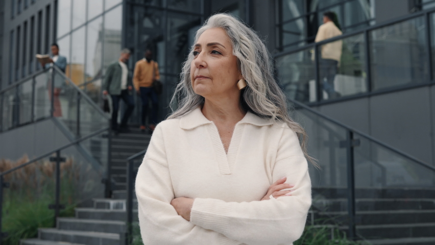 Charming aged business lady keeping arms crossed while standing at urban area and looking at camera. Portrait of successful and confident caucasian woman with grey hair.