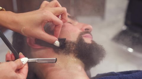 Barber shaving bearded man with sharp razor in Barber shop. Bearded male sitting in armchair in barbershop while hairdresser shaves his head with dangerous razor. High quality 4k footage