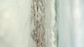 Vertical footage of the surf. Seashell beach, small seashells close-up. Sea foam from the surf, waves rolling along the shore. Beach vacation concept. Selective focus. 4K video, place for text
