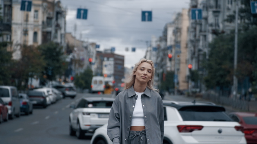 Attractive girl walking around city landscape with blonde hair on highway road. Confident female person smiling while going towards camera on road with passing cars. Walking and city life concept.