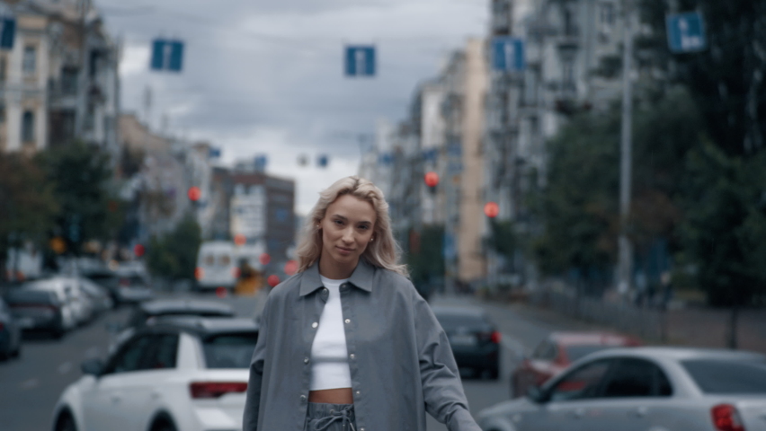 Attractive girl walking around city landscape with blonde hair on highway road. Confident female person smiling while going towards camera on road with passing cars. Walking and city life concept.
