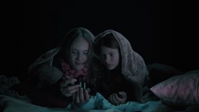Two Isolated Caucasian Girls Laying on the Floor Of a Dark and Cozy Bedroom on Top of Pillows and Blankets Watching a Video on Their Smartphone Device. Covered in Blankets, Laughing and Smiling.