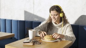Exited woman in yellow headphones have breakfast in cafe in sunny morning. Pretty joyful blonde lady travel blogger with stylish glasses eats bagel burger and dancing 