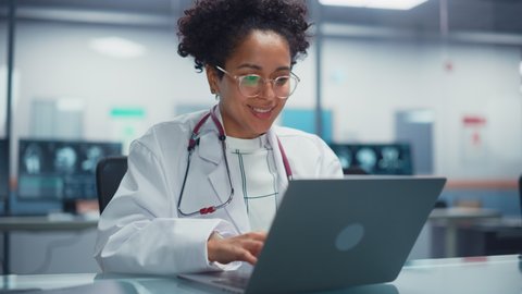 Hospital Doctors Office: Portrait of Young Promising Black Physician Works on Laptop Computer. Female Medical Professional in White Lab Coat Looking for Patient Treatment While Sitting at Her Desk