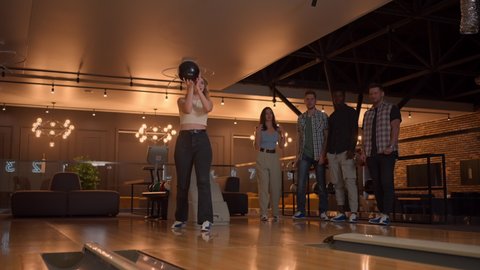 A young woman in bowling throws a ball on the track and knocks out a shot in slow motion and jumps and dances for joy. Friends fans support and clap with a smile.