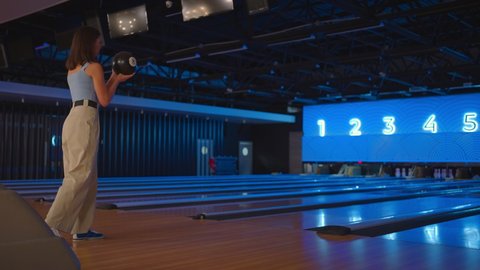A brunette woman in bowling throws a ball in slow motion and knocks down skittles. Play bowling. Throw balls on the floor of the bowling club