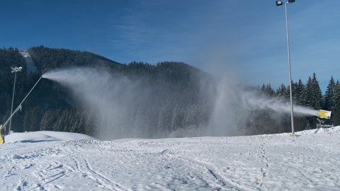 Snow cannon and snowgun make artificial snow on mountain slope on sunny morning at ski resort, backdrop of snowy spruce forest, ski track. Snow making system blowing artificial snow of water in winter