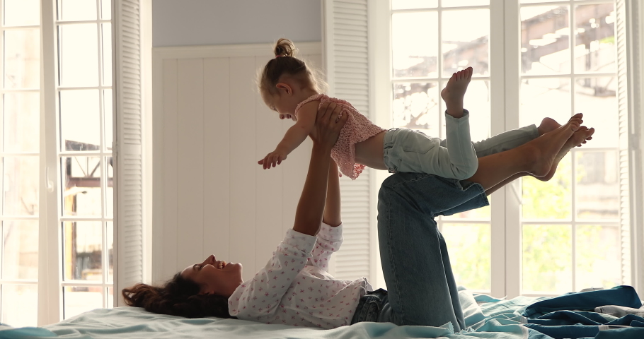 Woman lying on bed lifts her cute little 4s daughter on arms happy family spend leisure at modern house. Preschool girl spread arms like plane wings flying in air play with loving mom or nanny at home Royalty-Free Stock Footage #1085132198