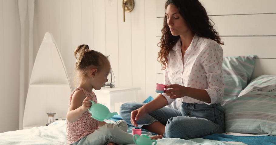 Young Hispanic mother her little daughter holds toy teacups plastic dish enjoy tea ceremony play together sit on bed in warm cozy bedroom. Weekend leisure activity with children, playtime, fun concept Royalty-Free Stock Footage #1085132216