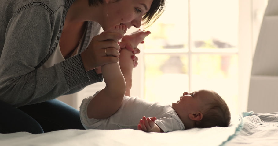 Loving mom kisses tiny toes of her cute 0-6 baby girl, close up. Smiling infant lying on bed enjoy mum caress and playtime looks satisfied. Babyhood, happy motherhood, mothers sincere love concept Royalty-Free Stock Footage #1085132297