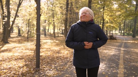 Close-up of aged gray-haired female suffering from heart burn during running in autumn park. Elderly woman jogger feeling pain during active cardio training and getting a heart attack