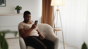 Excited African American Woman Reading Message On Smartphone Texting Sitting In Chair At Home. Great News Concept. Joyful Female Using Cellphone Receiving Positive Sms Communicating Online