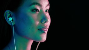 Attractive Young Asian Woman Illuminated By Neon Lights Listening Music In Earphones, Closeup Of Beautiful Korean Female Enjoying Favorite Songs While Standing Under Blue Green-Lighting, Slow Motion