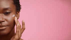 Facial Skincare. Half-Portrait Of Happy Black Woman Applying Moisturizer Cream On Face Smiling Looking At Camera Posing Over Pink Studio Background. Cropped Shot, Free Space For Text