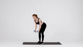 Young Sporty Female Doing Dumbbell Bent Over Fly Exercise In Studio, Athletic Millennial Woman Training Muscles While Standing On Fitness Mat Over White Background, Slow Motion Footage