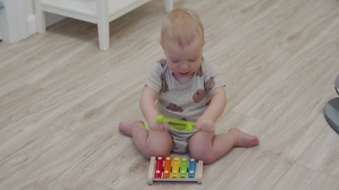 Cute little child sitting on the floor playing with colorful wooden xylophone musical toy for kids, 10 month old baby boy at home. Musical instruments for children to learn to play music. 