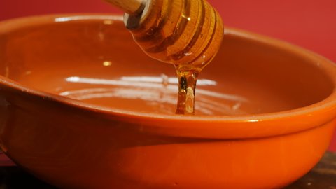Natural honey flows into a clay red pot from a honey spoon in a thin stream, forming a light glare from sunlight. New collection of fresh honey.