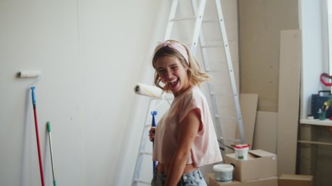 Woman singing and holding paint roller, female designer dancing, having fun. Happy housewife making overhaul, home renovating, painting walls. Smiling young wife during major maintenance. 