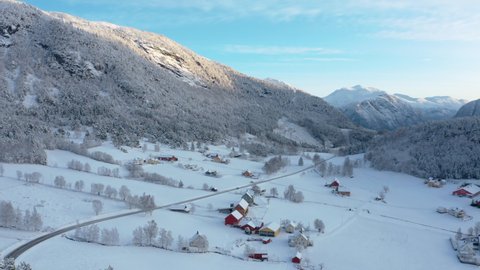 Liabygda village, hidden away on a snow covered Norwegian mountain ledge. Aerial shot, slow pan revealing the tranquil and secluded cold frosty farm land. Low sun slightly brushing the mountain peaks.
