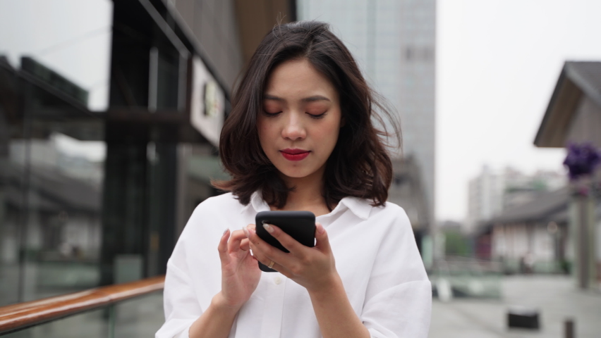 Happy young pretty asian woman holding mobile phone looking browsing online urban people typing on cellphone walking in the city Royalty-Free Stock Footage #1085160860