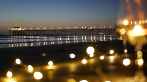 Candle flame lights in glass, romantic beach date, California ocean waves, sea water. Candlelight. Wineglass on sand, garland in twilight dusk. Illuminated pier reflection. Seamless looped cinemagraph
