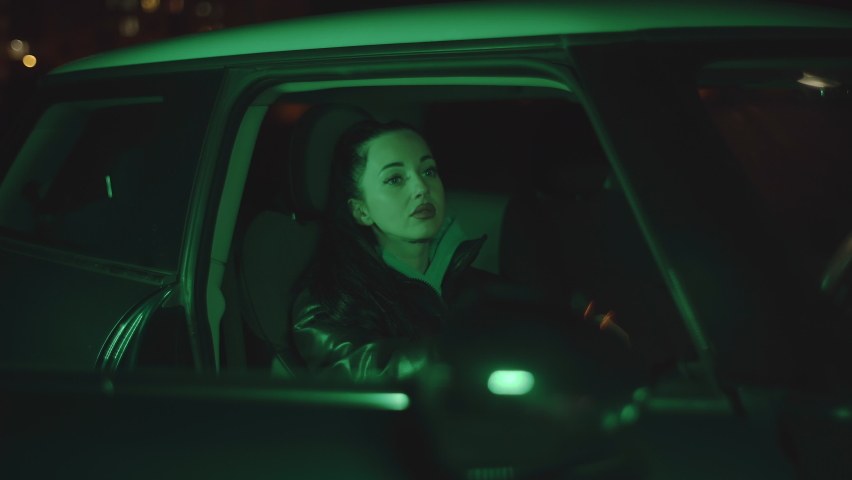 Pretty brunette girl sitting in car at dark night. Beautiful woman passenger looking out of window in car in urban city street in neon green light Royalty-Free Stock Footage #1085165153