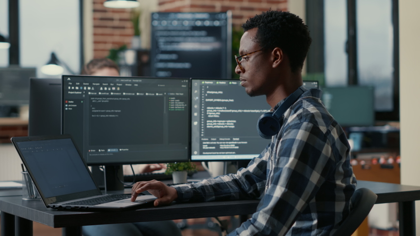 Portrait of african american developer using laptop to write code sitting at desk with multiple screens parsing algorithm in software agency. Coder working on user interface using portable computer. Royalty-Free Stock Footage #1085169626