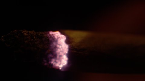 MACRO, DOF: Detailed shot of a burning joint of a person smoking weed at night. Close up view of a cigarette burning as smoker takes a long drag in the darkness. Tobacco and rolling paper burning