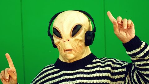 Ufo Alien mask with human clothes dance and listen music song with headphones and green wall in background - concept of earth planet and extraterrestrial in living between us