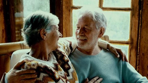 Old people senior man and woman in love and tenderness at home. Mature elderly couple enjoy relationship hugging and caring each other sitting on the sofa in living room with outdoor view