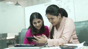 young Indian two woman are laughing while watching a funny video on a smartphone or cellphone sitting at office room.Two happy friends watching a movie on a mobile phone.