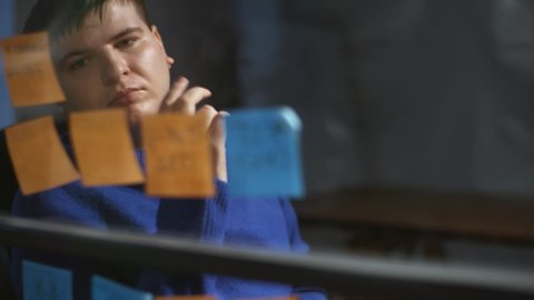 Chest-up of young Caucasian man wearing blue long sleeve pullover, writing on sticky note on glass wall in office in evening